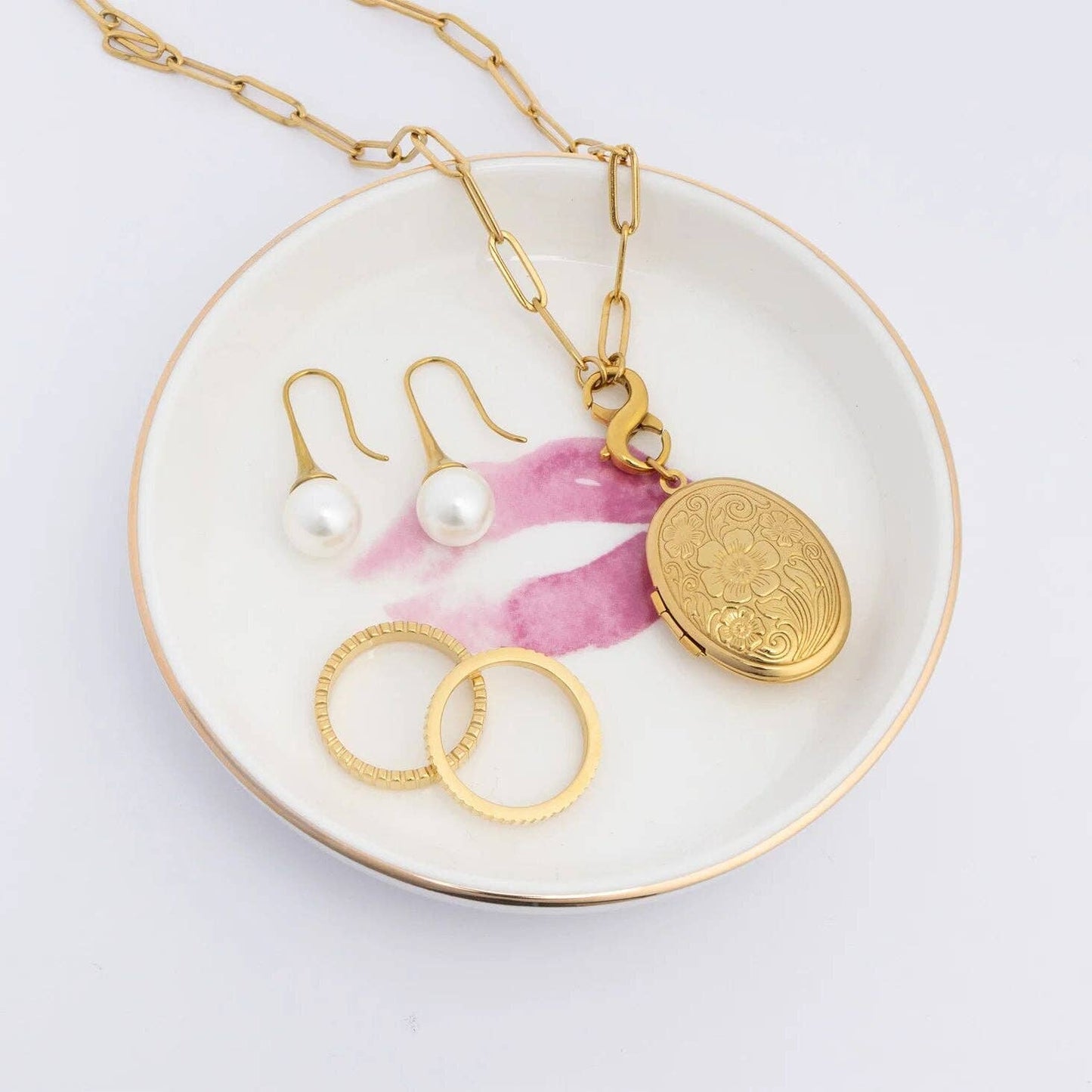 Gold necklace with a pendant, pearl earrings, and gold rings on a decorative plate with floral design.