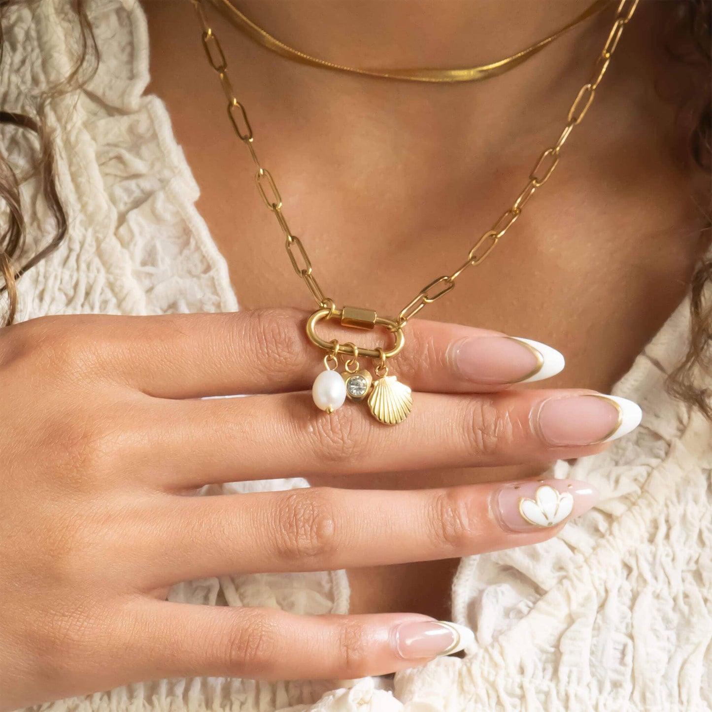 Close-up of a hand wearing a gold necklace with shell and pearl charms, against a neutral background.