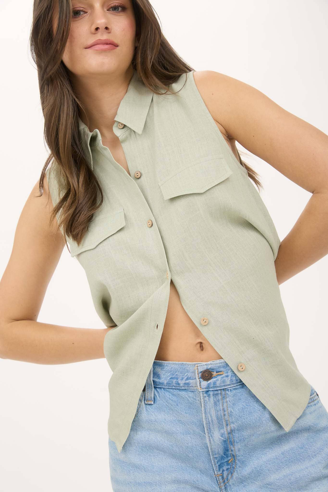 Woman wearing a light green sleeveless button-up shirt with denim shorts on a white background