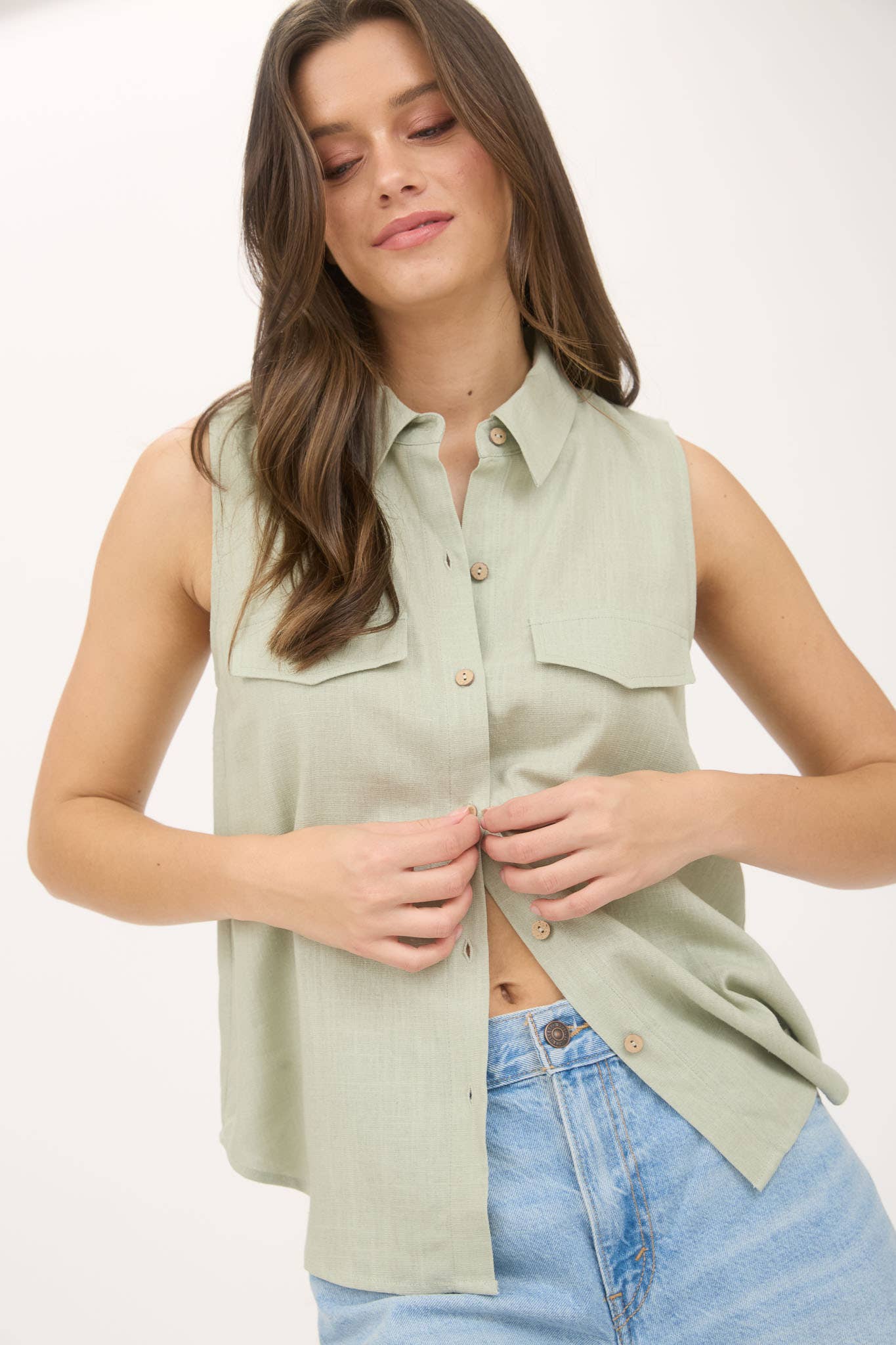 Woman wearing a light green sleeveless button-up shirt with blue jeans on a white background