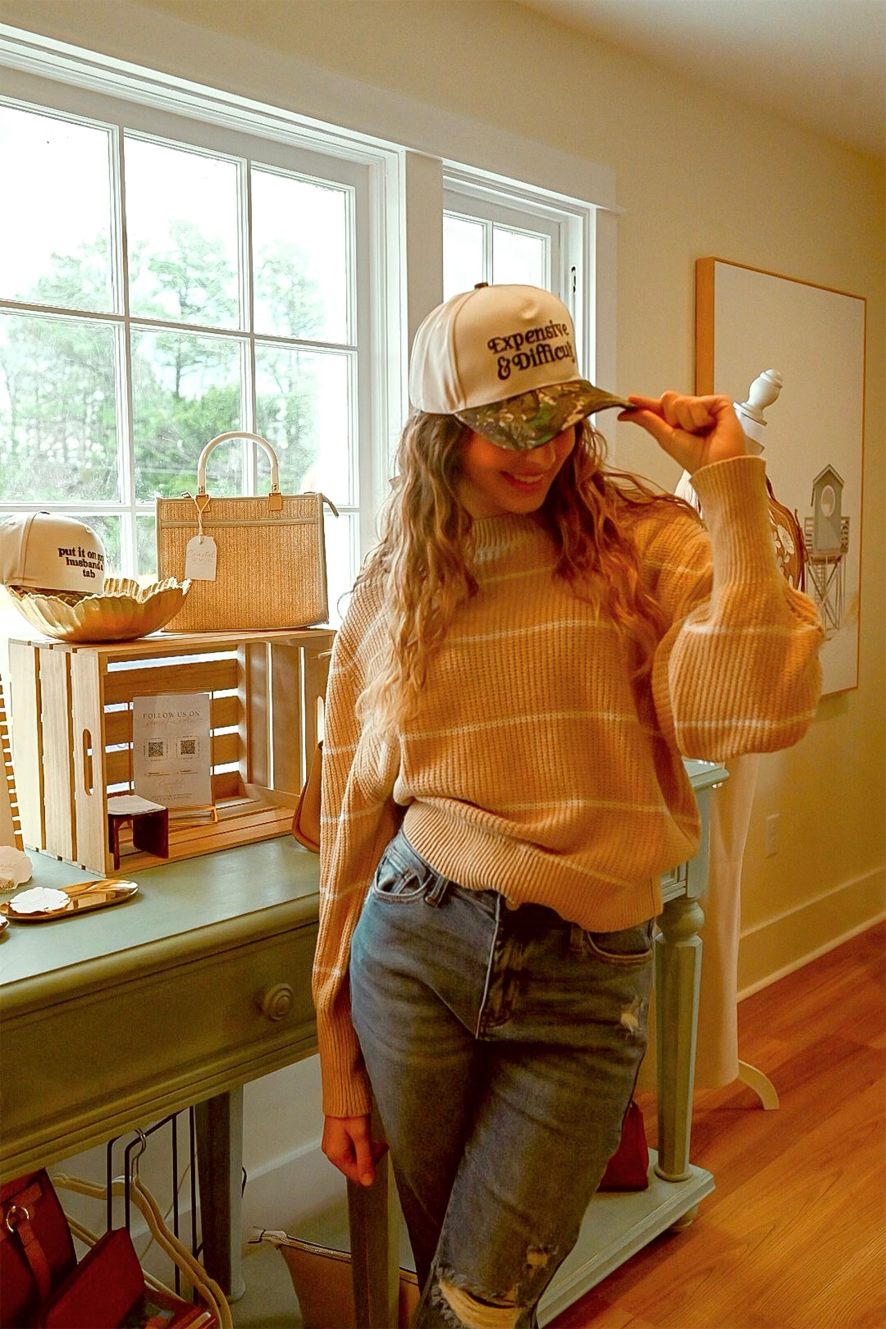 Person wearing a beige sweater and cap indoors, with a window and furniture in the background.