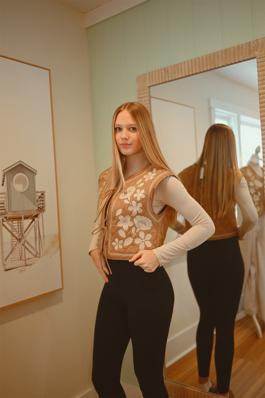 Woman standing in front of a mirror wearing a patterned vest and black pants, with a framed picture on the wall.