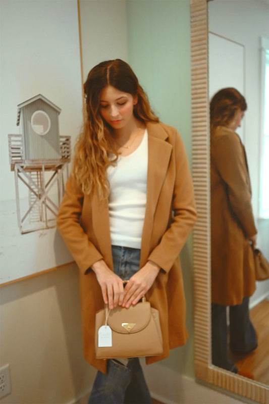 Woman holding a beige handbag in front of a mirror, wearing a brown coat.