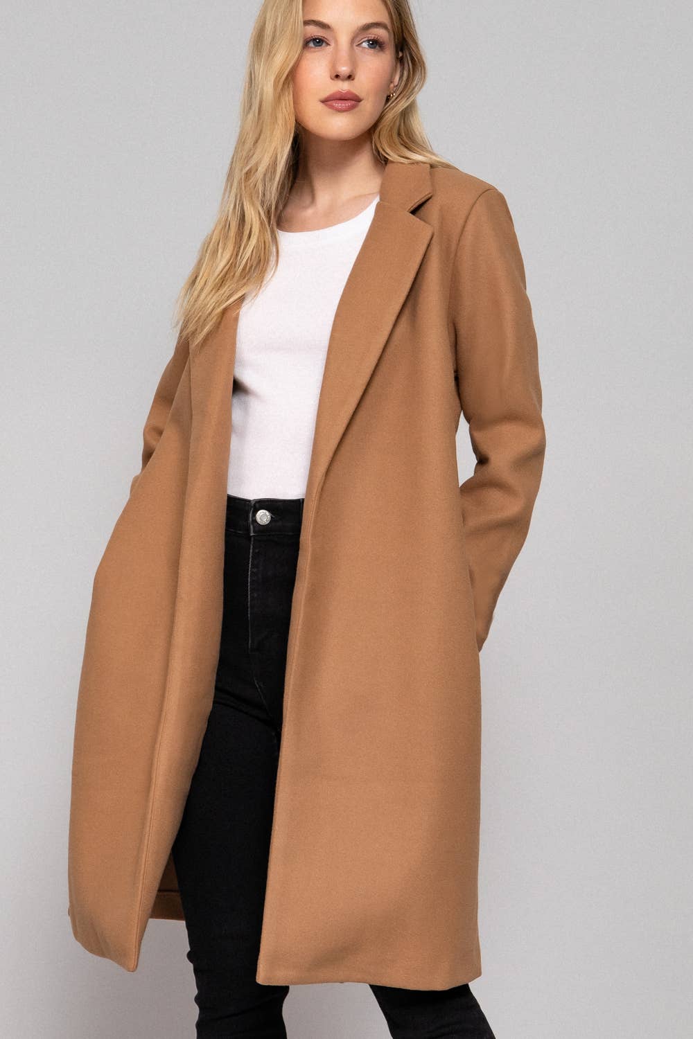 Woman wearing a long brown coat over a white top and black pants on a light gray background