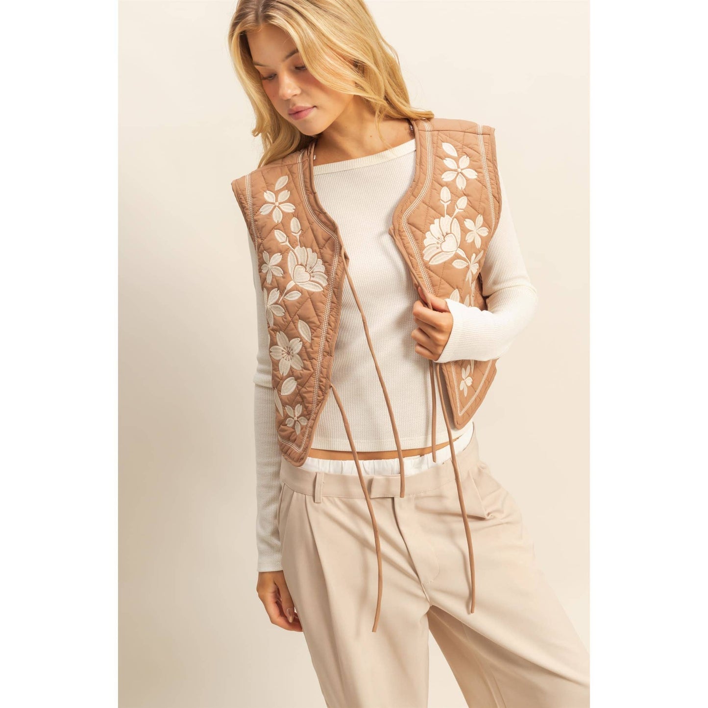 Woman wearing a beige floral embroidered vest over a white top and beige pants on a light background