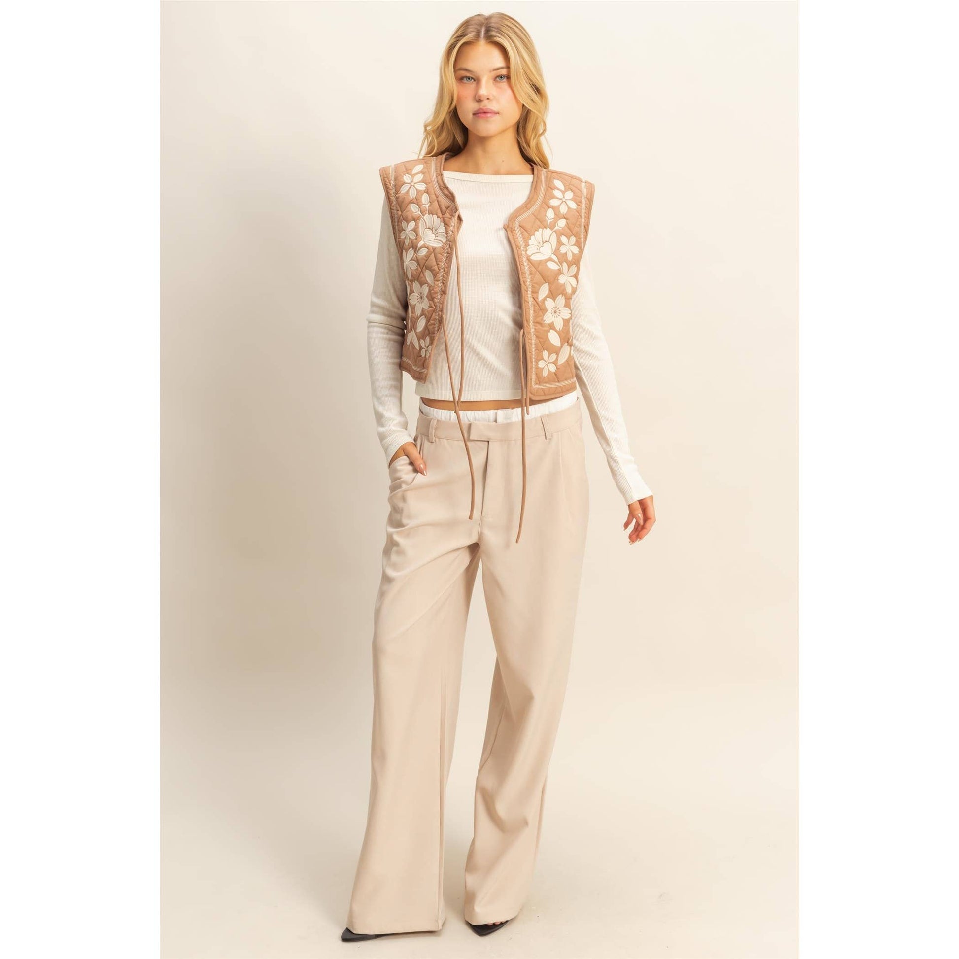 Woman wearing a beige floral vest over a white top and beige pants on a light background