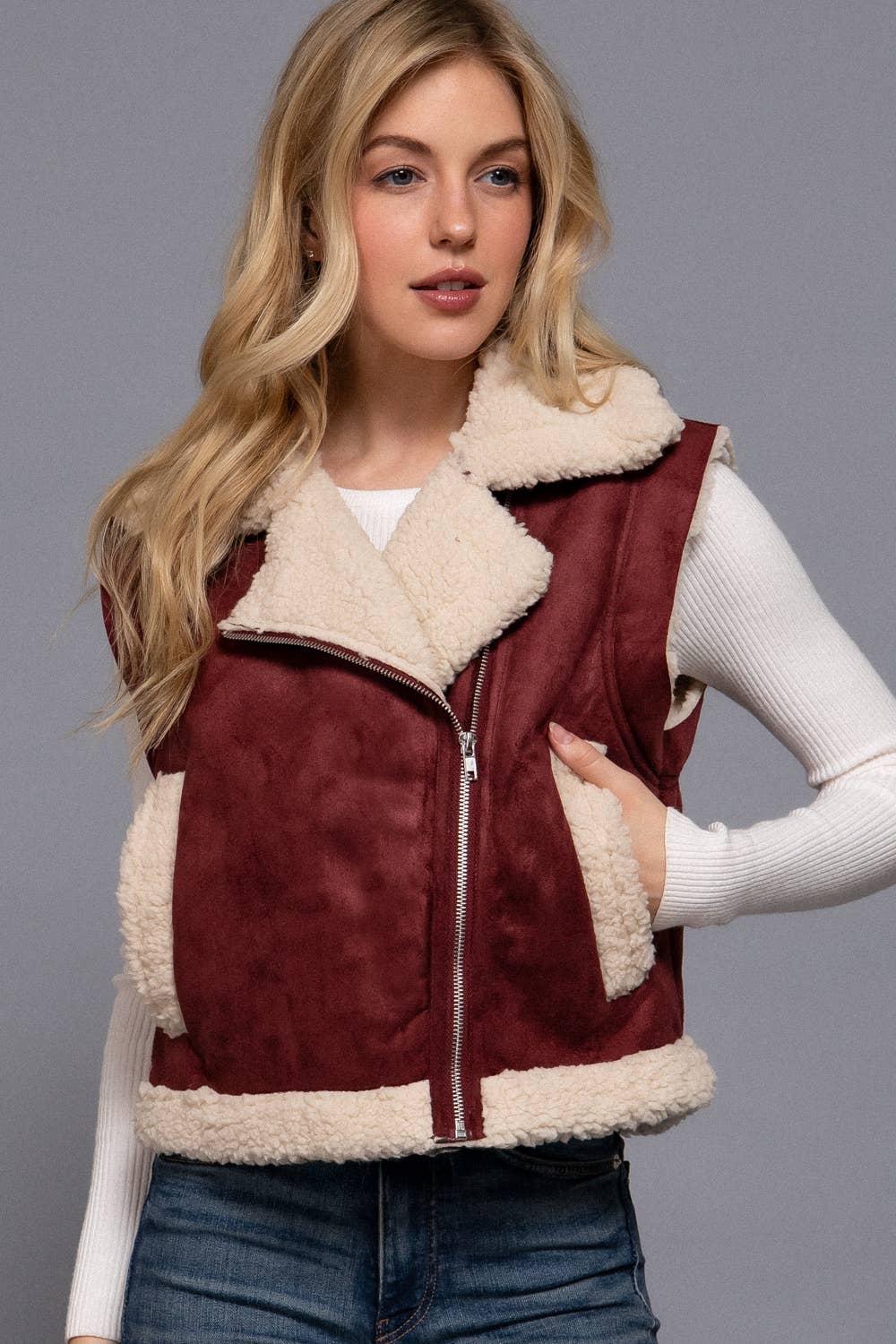 Woman wearing a burgundy suede vest with white fur lining on a gray background